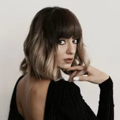 Kobieta z modnym, **blunt bob** w odcieniach brązu i blondu, z grzywką, w czarnej sukience z odkrytymi plecami.
