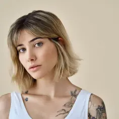 Młoda kobieta z blond włosami, w białej bluzce, z tatuażami na ramieniu i klatce piersiowej. Jej fryzura to modny clavi cut.