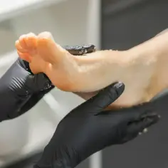 Dłonie w czarnych rękawiczkach wykonują peeling stopy. To część zabiegu, który nazywa się pedicure.