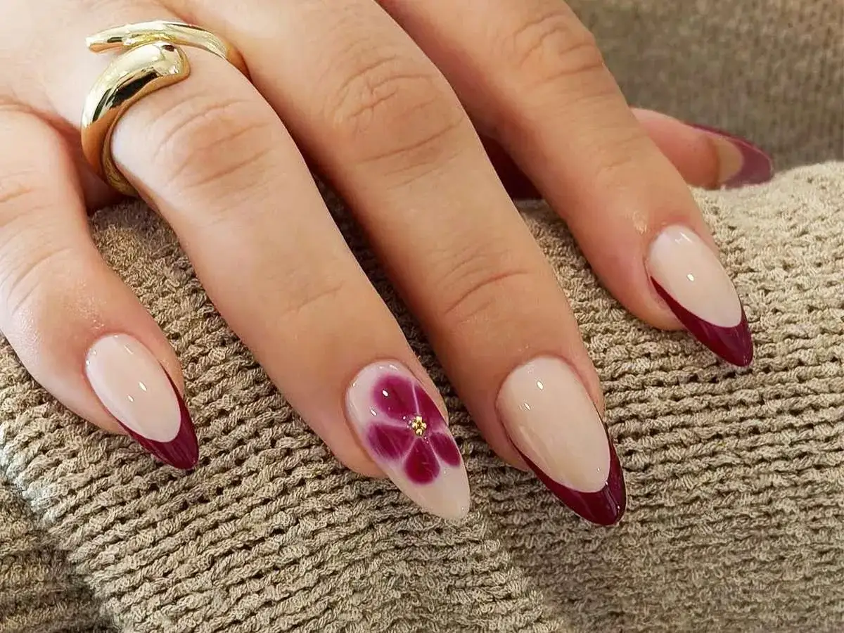 Elegancki manicure francuski z bordowymi końcówkami i kwiatowym zdobieniem na jednym paznokciu. Złoty pierścionek dodaje blasku.