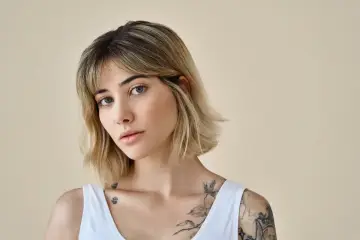 Młoda kobieta z blond włosami, w białej bluzce, z tatuażami na ramieniu i klatce piersiowej. Jej fryzura to modny clavi cut.