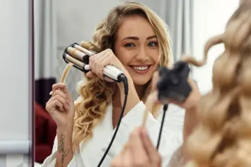 Uśmiechnięta blondynka tworzy piękne fale na włosach za pomocą lokówki.