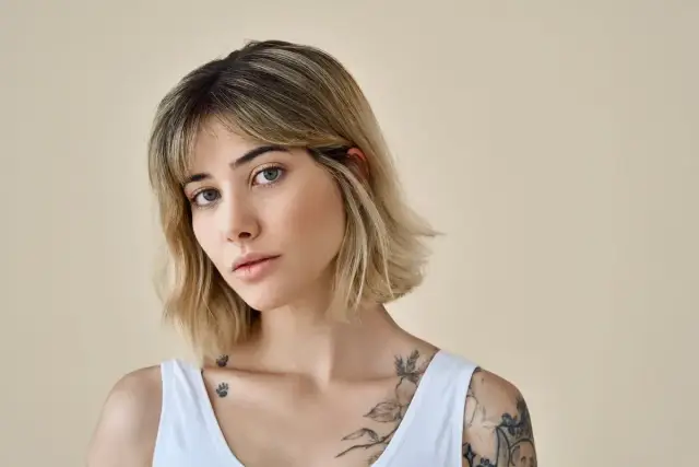 Młoda kobieta z blond włosami, w białej bluzce, z tatuażami na ramieniu i klatce piersiowej. Jej fryzura to modny clavi cut.